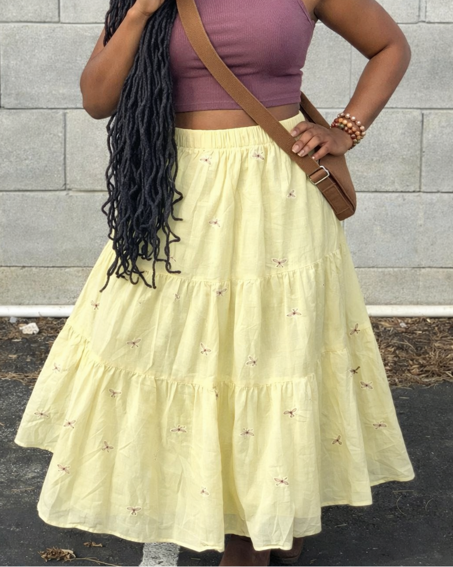 Woman wearing a purple top and yellow skirt with a brown bag against a gray brick wall.
