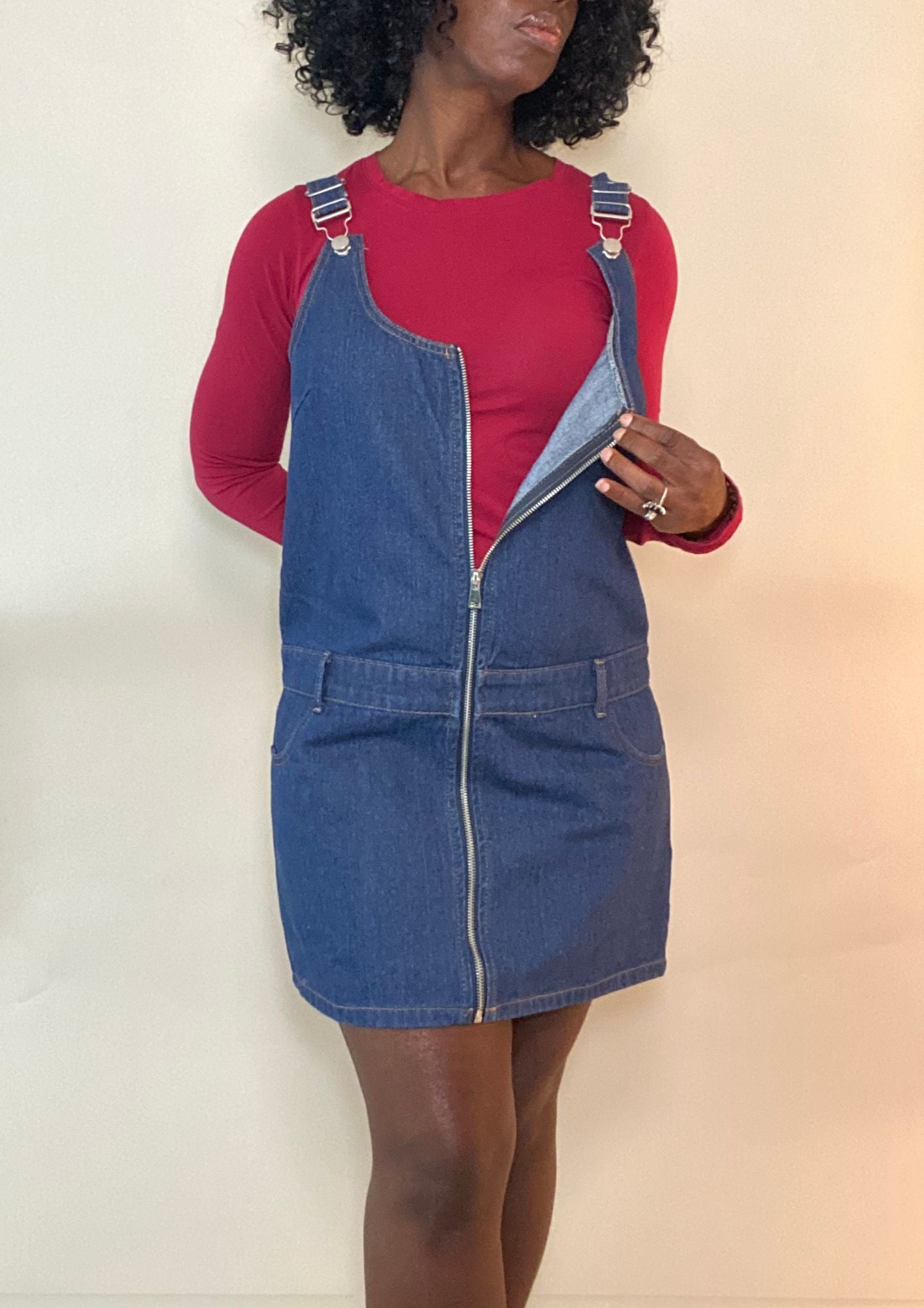 Women’s zip-front denim overall mini dress over a red long-sleeve top, partially unzipped — fall women’s denim dress outfit idea.