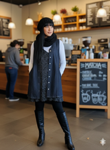 Woman standing in a coffee shop wearing a dark vest and black boots.
