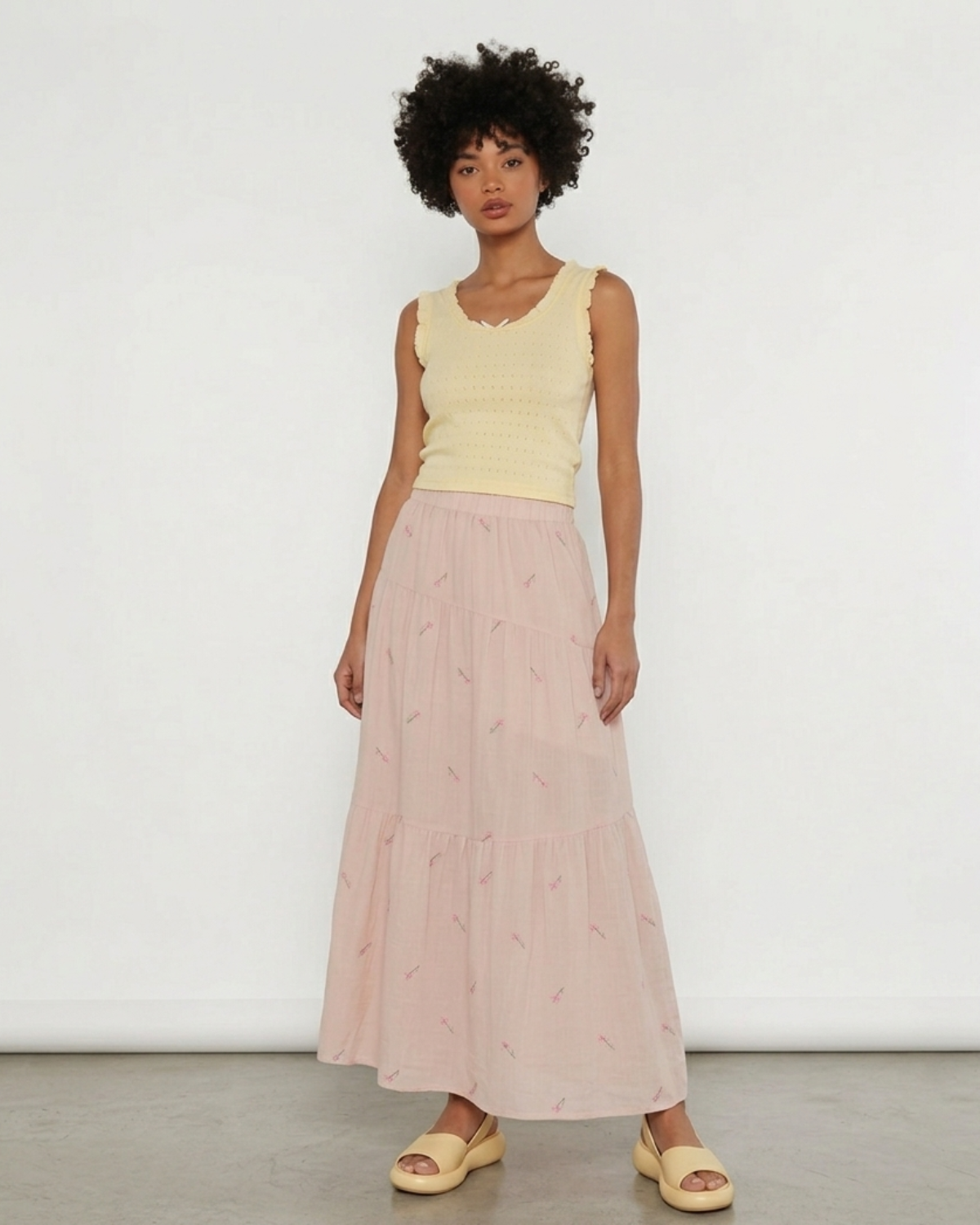 Woman wearing a blush pink floral embroidered tiered midi skirt with a light yellow sleeveless top.