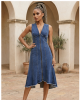 Woman wearing a long denim dress in classic blue with panel seams, front zip detail, sleeveless V-neck design, and raw hem, standing in a courtyard with arches and potted plants.