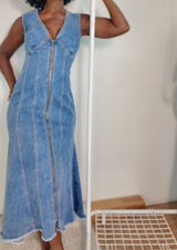 Close-up front of long denim zip-front dress in classic denim blue showing V-neck, paneled seams, and raw hem.