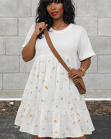 Woman wearing a white dress with floral patterns against a gray brick wall.